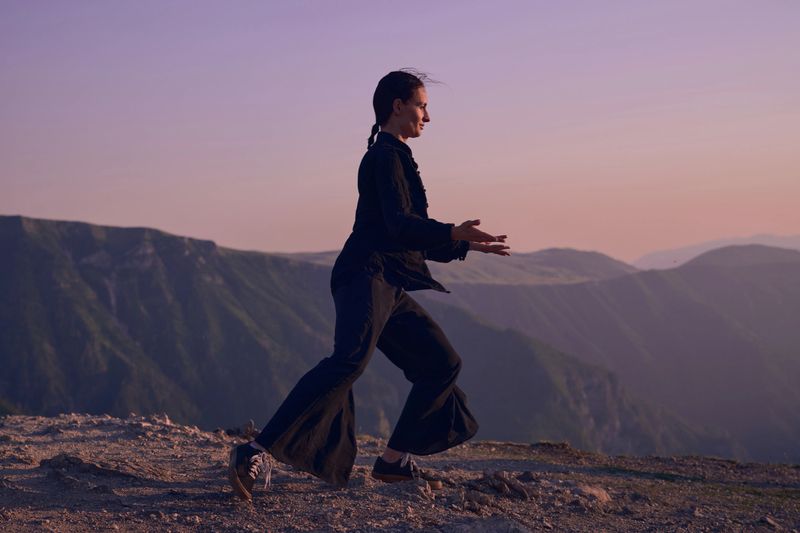 A woman practicing Tai Chi in a traditional black outfit on a mountain at sunrise, her arms outstretched in a meditation pose, focuses on balance, energy, and mindfulness in nature.