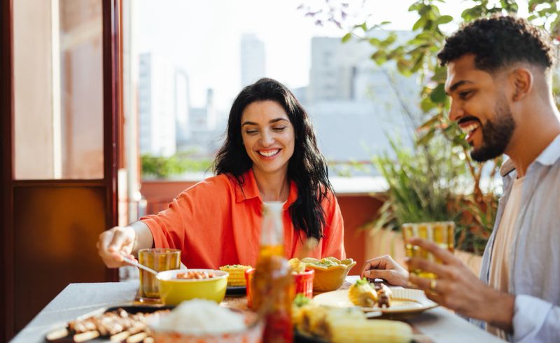 Two joyful Latin American individuals enjoying a shared meal of local food on a terrace with city views.