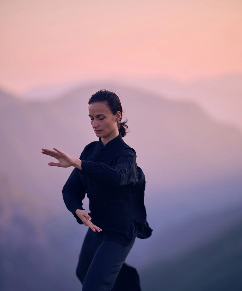 A woman practicing Tai Chi in a traditional black outfit on a mountain at sunrise