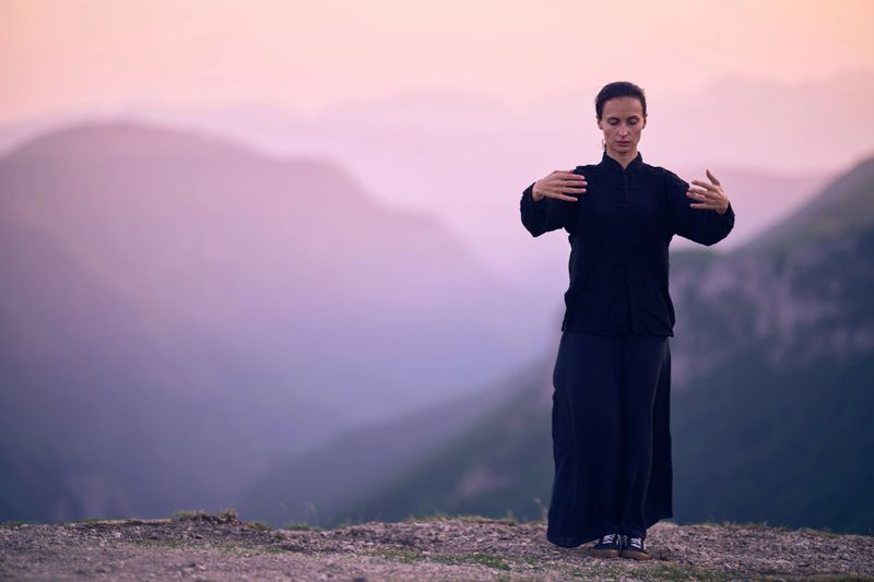 A woman practicing Tai Chi in a traditional black outfit on a mountain at sunrise, her arms outstretched in a meditation pose, focuses on balance, energy, and mindfulness in nature.