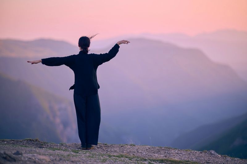 A woman practicing Tai Chi in a traditional black outfit on a mountain at sunrise, her arms outstretched in a meditation pose, focuses on balance, energy, and mindfulness in nature.