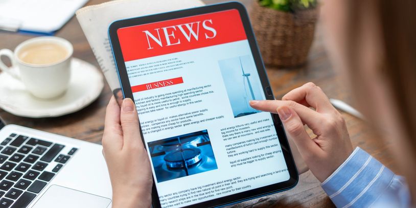 Person reading business news on a tablet at a desk with coffee and laptop.