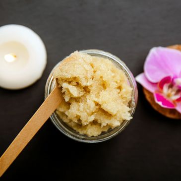 A jar of sugar scrub with a wooden spatula beside a candle and orchid flower.
