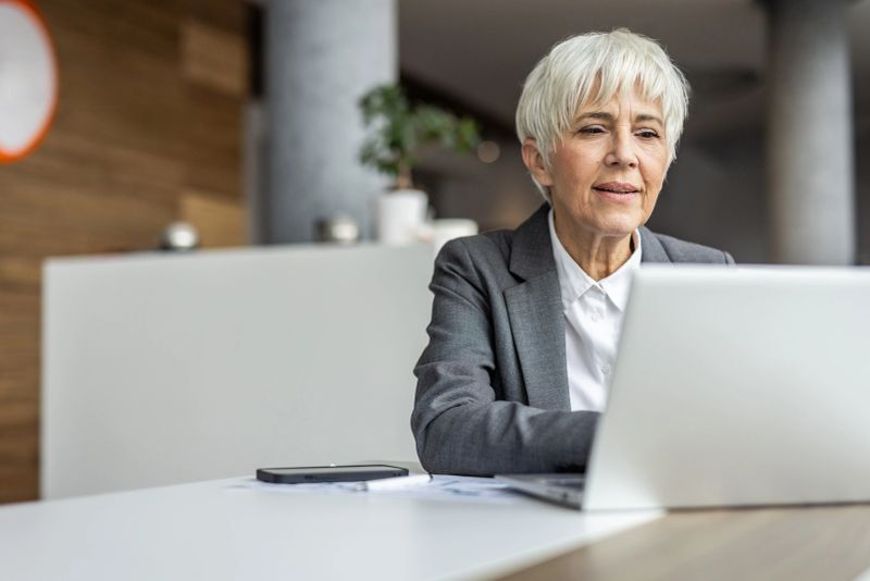 Focused mature businesswoman concentrating on her work, using laptop at her desk in a bright, modern office environment, demonstrating professionalism and expertise