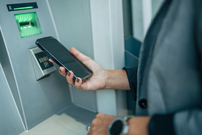Businesswoman withdrawing cash from an atm while using her smartphone for a seamless contactless transaction, showcasing the convenience of modern digital banking in an urban setting