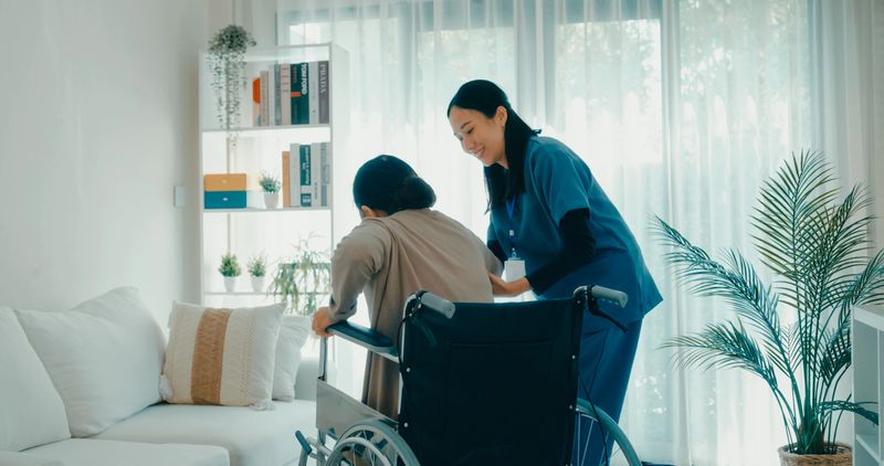 Young Asian nurse helps an elderly woman in a wheelchair support and care in living room at home. Home health care concept.