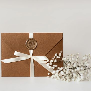 Elegant brown envelope sealed with wax and tied with a white ribbon, next to delicate white flowers.
