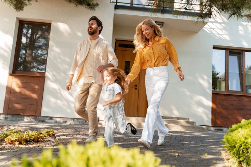 Cheerful parents and daughter running out of their modern house holding hands, enjoying sunny day together, expressing joy and family unity. Parenthood concept