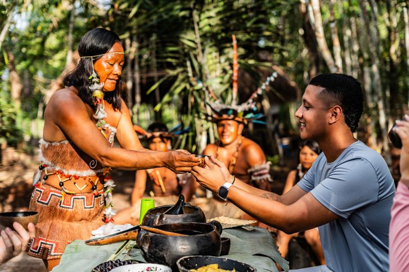 Curripaco community member offers traditional hospitality serving typical foods to group of visitors in natural village environment, demonstrating cultural generosity and authentic gastronomic exchange through respectful ethnic tourism in the Amazonian region