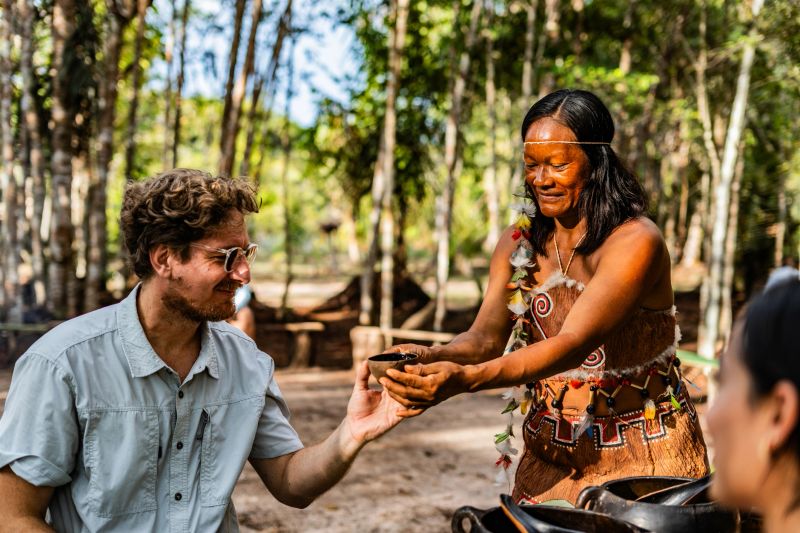 Curripaco community member offers traditional hospitality serving typical foods to group of visitors in natural village environment, demonstrating cultural generosity and authentic gastronomic exchange through respectful ethnic tourism in the Amazonian region