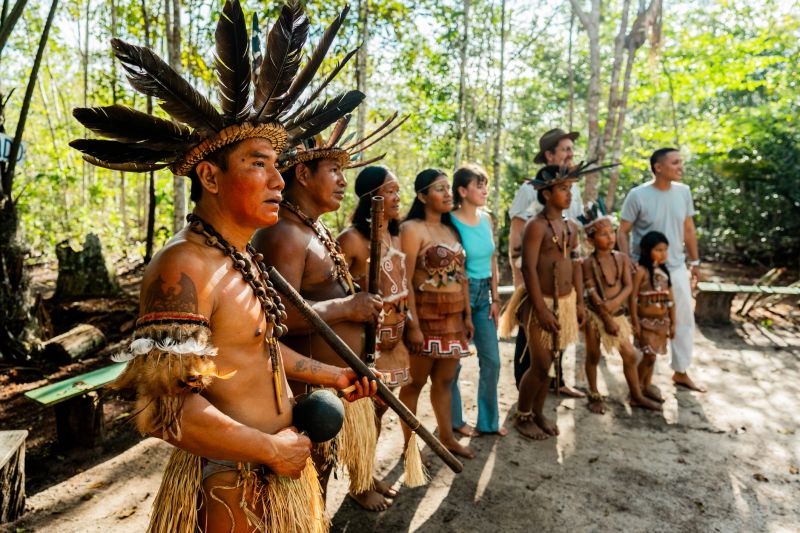 Curripaco community members wearing traditional ornaments and headdresses pose alongside visitors in natural village environment, demonstrating respectful cultural exchange and harmonious coexistence that promotes appreciation of ethnic diversity through sustainable tourism in the Amazonian region