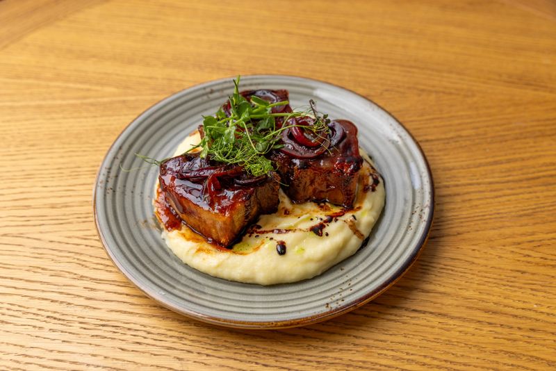 Glazed pork belly cubes served on a bed of creamy mashed potatoes, garnished with caramelized red onions and fresh microgreens, elegantly plated on a gray ceramic dish over a wooden table