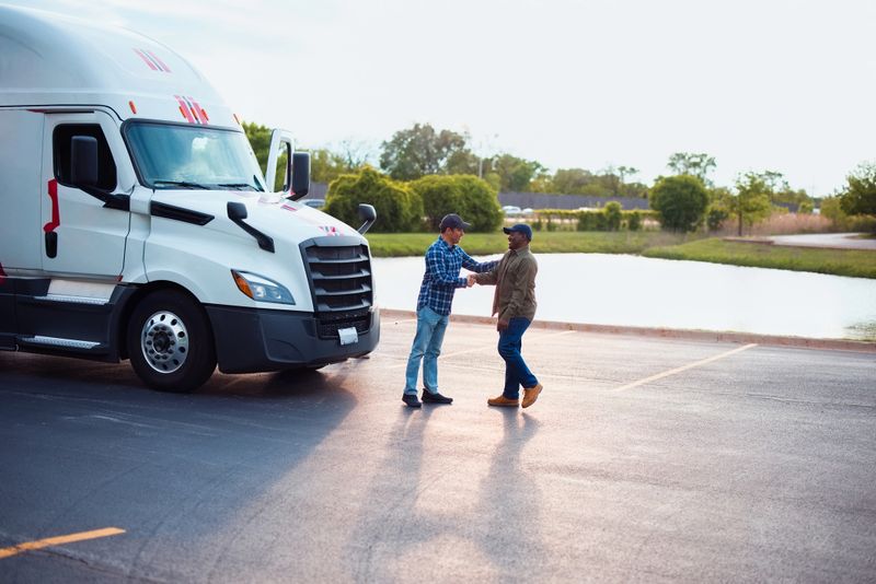 Two truck drivers meeting and shaking hands in a parking area next to a large truck, with a serene outdoor environment in the background, symbolizing teamwork and collaboration within the logistics industry.