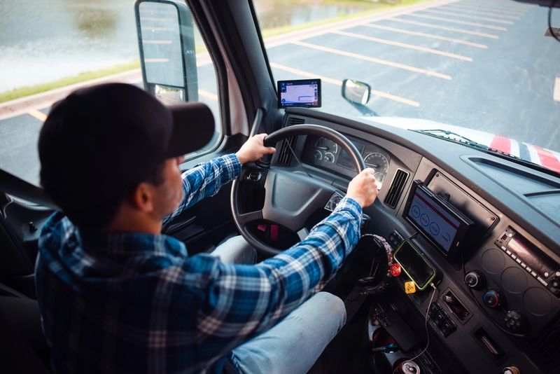 A truck driver steering a vehicle while monitoring navigation systems and the road ahead, emphasizing professional driving practices and technology.