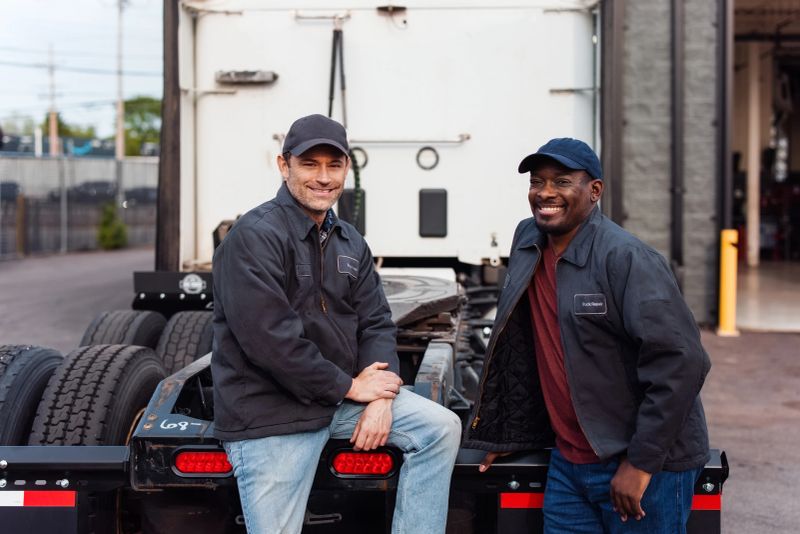 Two smiling mechanics stand outside in a workshop setting posing by a semi-truck trailer. Both wear uniforms and caps, evoking a professional but friendly working atmosphere, representing teamwork and professionalism.