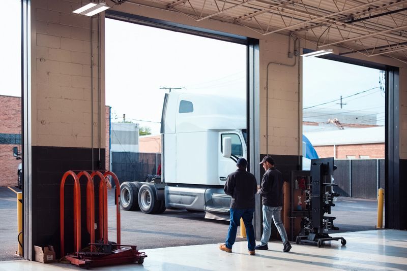 Two workers stand in a garage, observing a parked semi-truck. The scene depicts a moment of vehicle inspection or maintenance in an industrial setting.