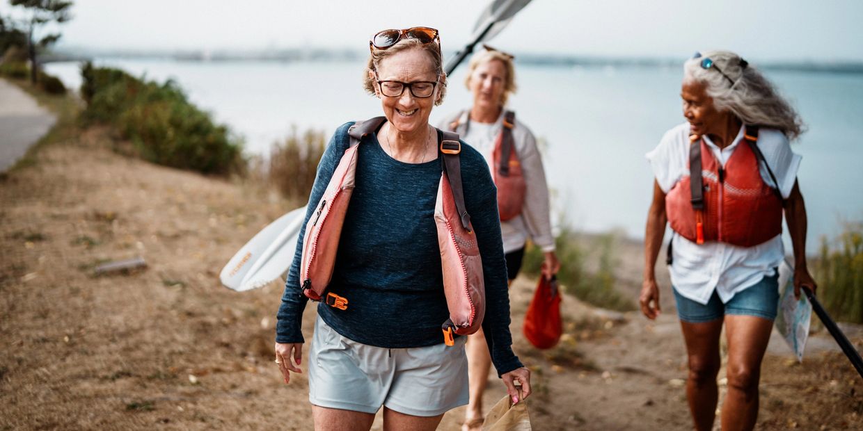 Three women enjoy paddling by the water with life vests on.