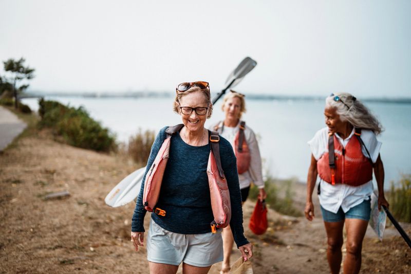 A smiling senior woman walks up a sandy path as her sister and their friend follow behind, after kayaking together in Casco Bay off the shore of Portland, Maine.