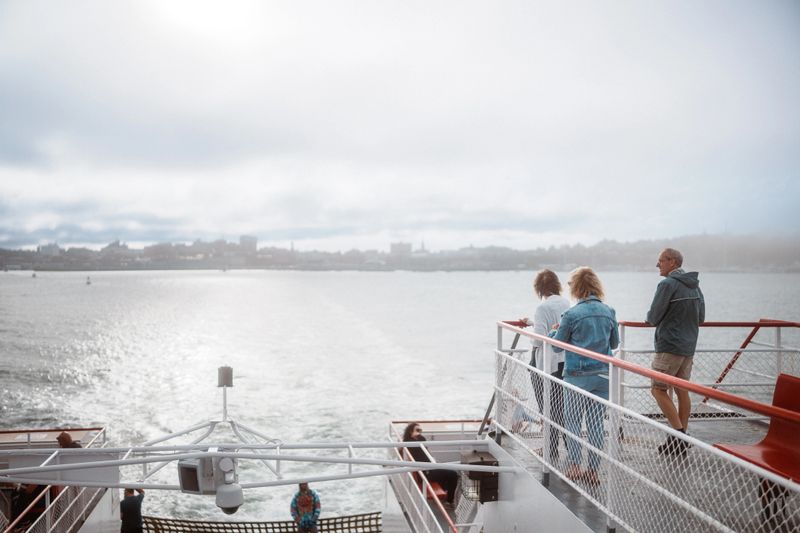 Three senior friends stand on the deck of a ferry looking back at the Portland, Maine skyline in the distance while traveling to Peaks Island.