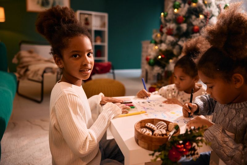 Three happy preschool age Black sisters are drawing and coloring pictures together at a small table in their cozy living room during the Christmas holiday season