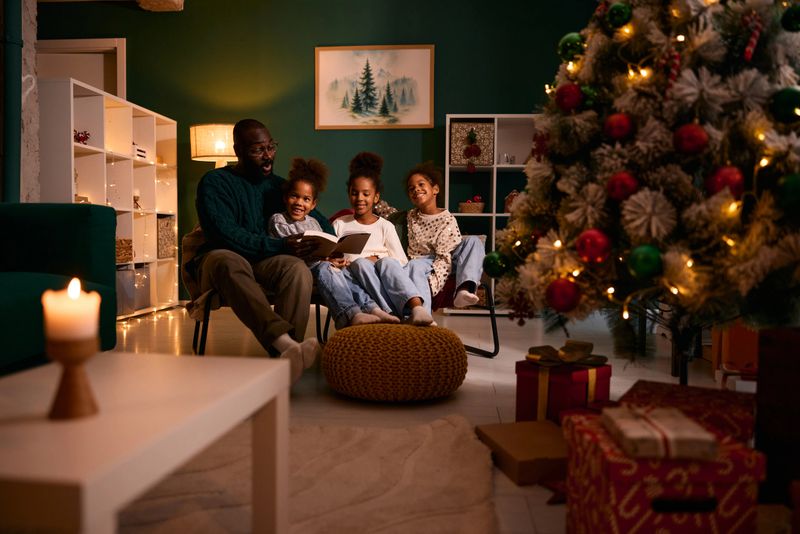 African American father sitting with his three children, two girls and one boy, reading a book together in a cozy living room decorated for Christmas with a lit tree and wrapped presents
