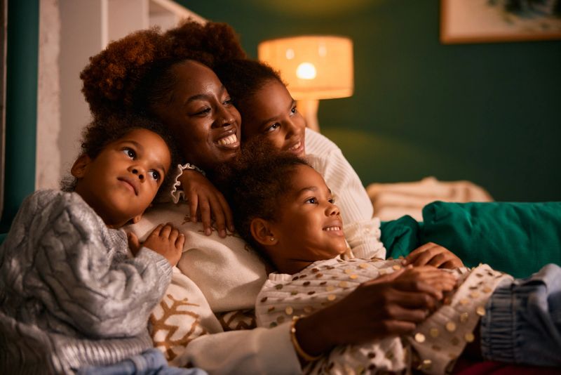 Black mother and her three young daughters relaxing on the couch in a cozy living room, enjoying a movie or show together, sharing a happy and loving family moment