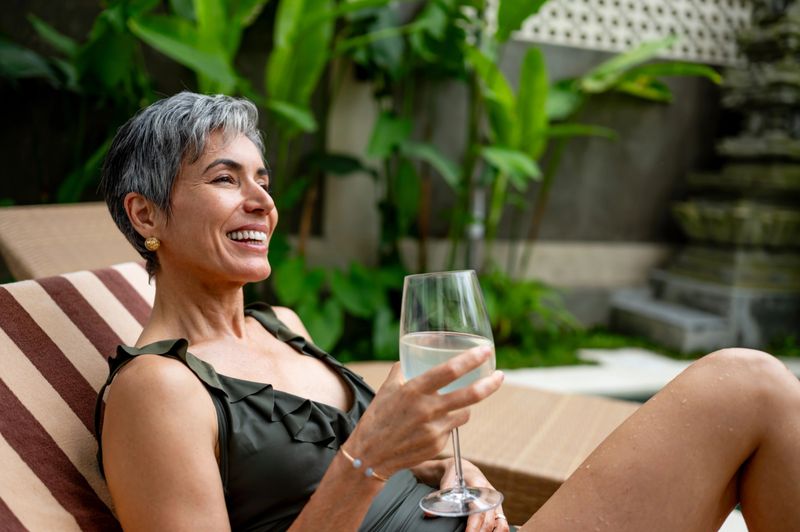 A cheerful woman with gray hair enjoys a beverage while lounging comfortably in a tropical setting. Surrounded by greenery, she wears a swimsuit, radiating relaxation, elegance, and vacation vibes.