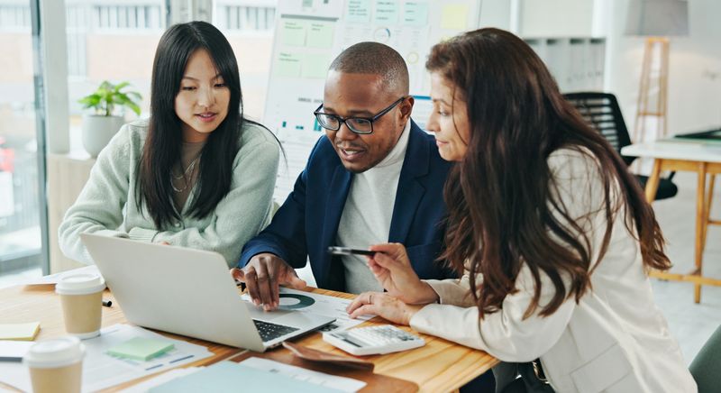 A group of professionals engaged in discussion and working on a project together with a laptop in a corporate office environment. This image depicts teamwork and collaboration in achieving work-related goals.