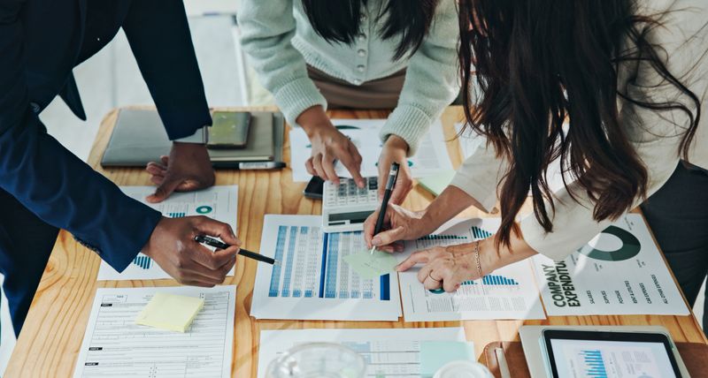 Diverse group of professionals collaborating on data analysis and financial planning at a table with documents, charts, and calculators, illustrating teamwork, productivity, and strategic business management.