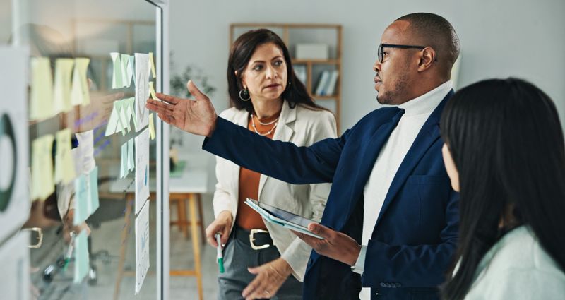 A business team collaborates at a glass board, discussing strategies and concepts in a modern office environment. Teamwork and brainstorming are evident as the group shares ideas and makes notes.
