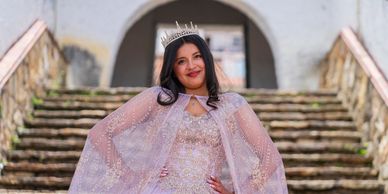 A young woman in a sparkling gown and crown poses confidently on stone steps.