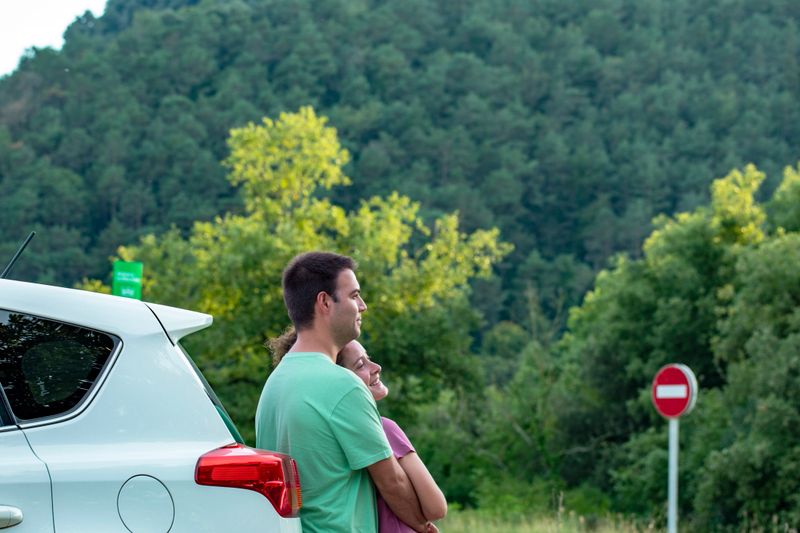 A couple shares a tender moment, hugging near their car in a peaceful natural setting surrounded by trees and greenery.