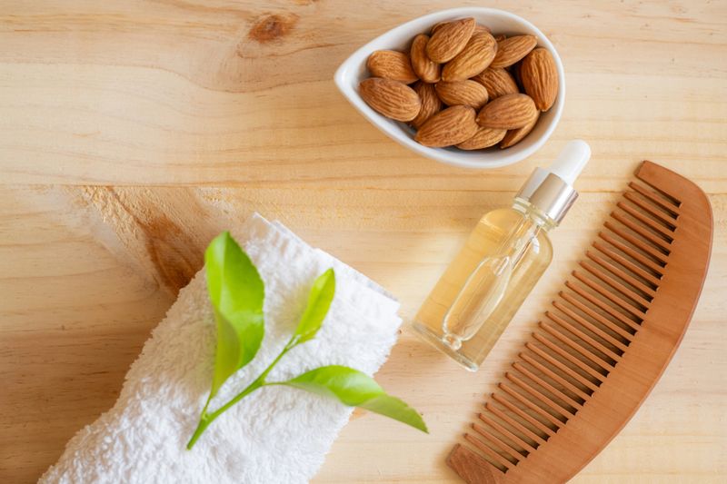 Almond oil dropper bottle, wooden comb, white towel, and fresh almonds arranged for a natural hair care routine, emphasizing shine, strength, and hydration