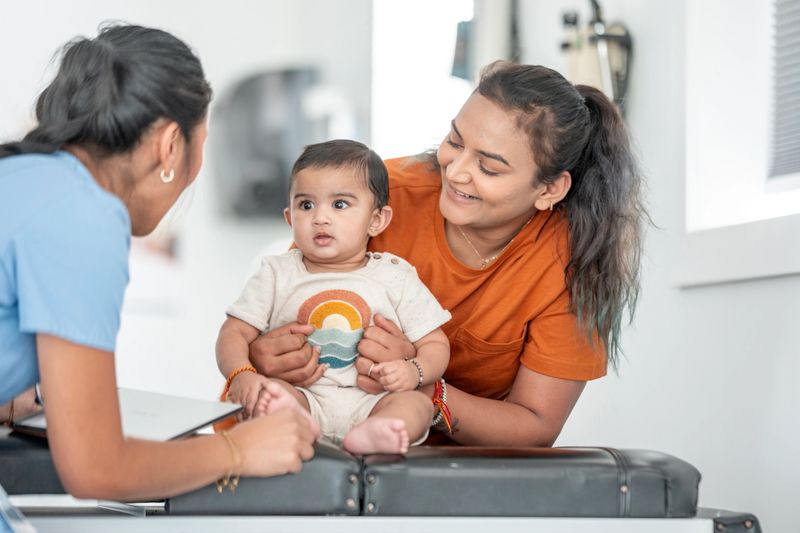 A cheerful mother holds her baby as a caring pediatrician interacts with them during a routine medical checkup at a clinic. The scene portrays compassion, health, and positive human connection.