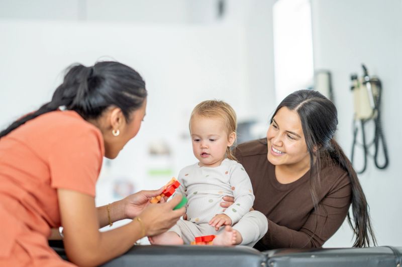 An attentive Asian nurse engages a baby during a consultation as the parent watches. The interaction showcases care and connection in a clinical environment, promoting early childhood care and medical trust.