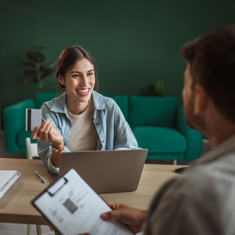 Woman holding a credit card and smiling at a man during a business meeting in a modern office, showing online payment, e-commerce, and financial transaction concepts