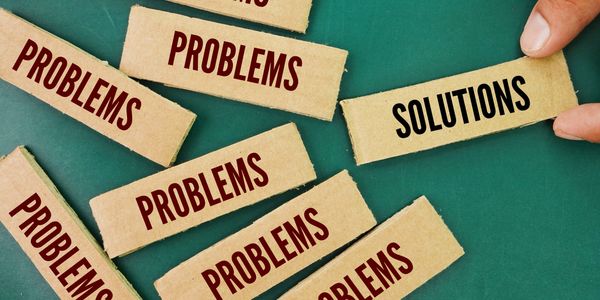 A hand holding a cardboard piece labeled 'SOLUTIONS' among many pieces labeled 'PROBLEMS'.