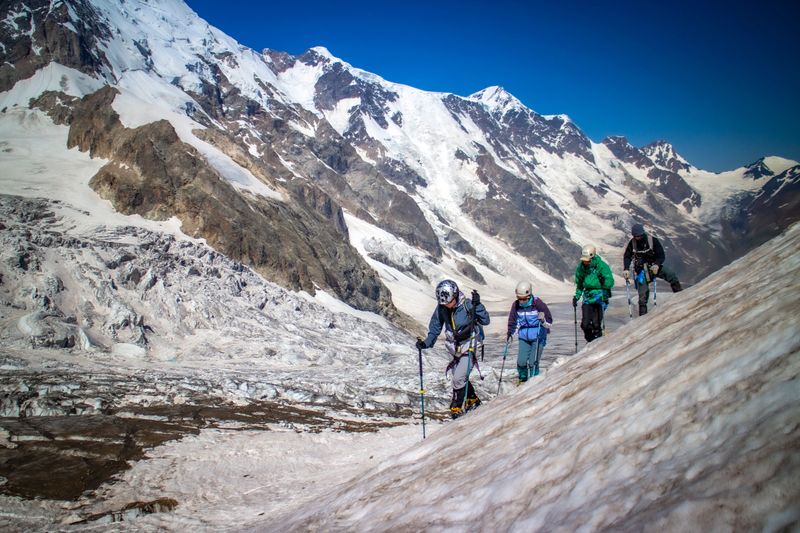 A group of mountain hikers makes their way across a steep glacier surrounded by dramatic alpine scenery. In the background rise towering snowy peaks and massive ice formations. The image reflects adventure, endurance, teamwork, and exploration in extreme high-altitude conditions.