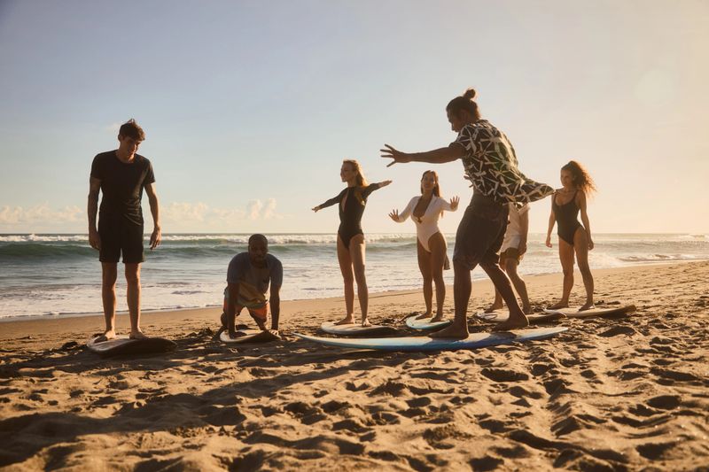 A group of individuals participates in a surfing lesson on a sandy beach, practicing techniques under a clear sky, enjoying the coastal atmosphere and fostering a spirit of learning and adventure.