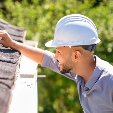 Construction worker inspecting roof shingles while wearing a white hard hat.