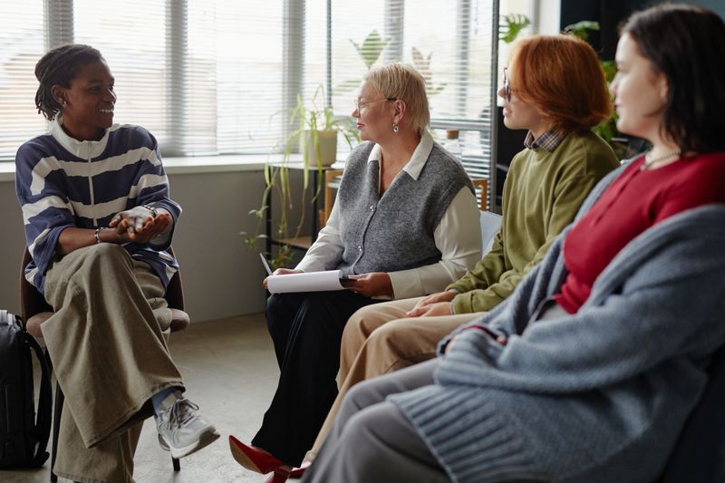 Group of multiethnic adults sitting in circle engaging in discussion, Black man gesturing while speaking, Caucasian middle aged woman holding notepad, two young adults listening attentively