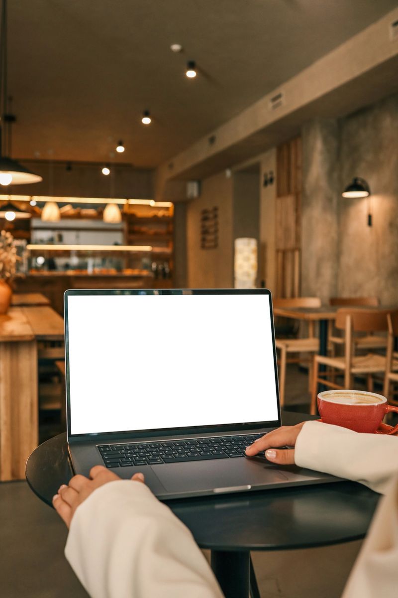 Person using laptop with blank screen at cafe table, red coffee cup nearby, cozy interior with warm lighting and wooden furniture.
