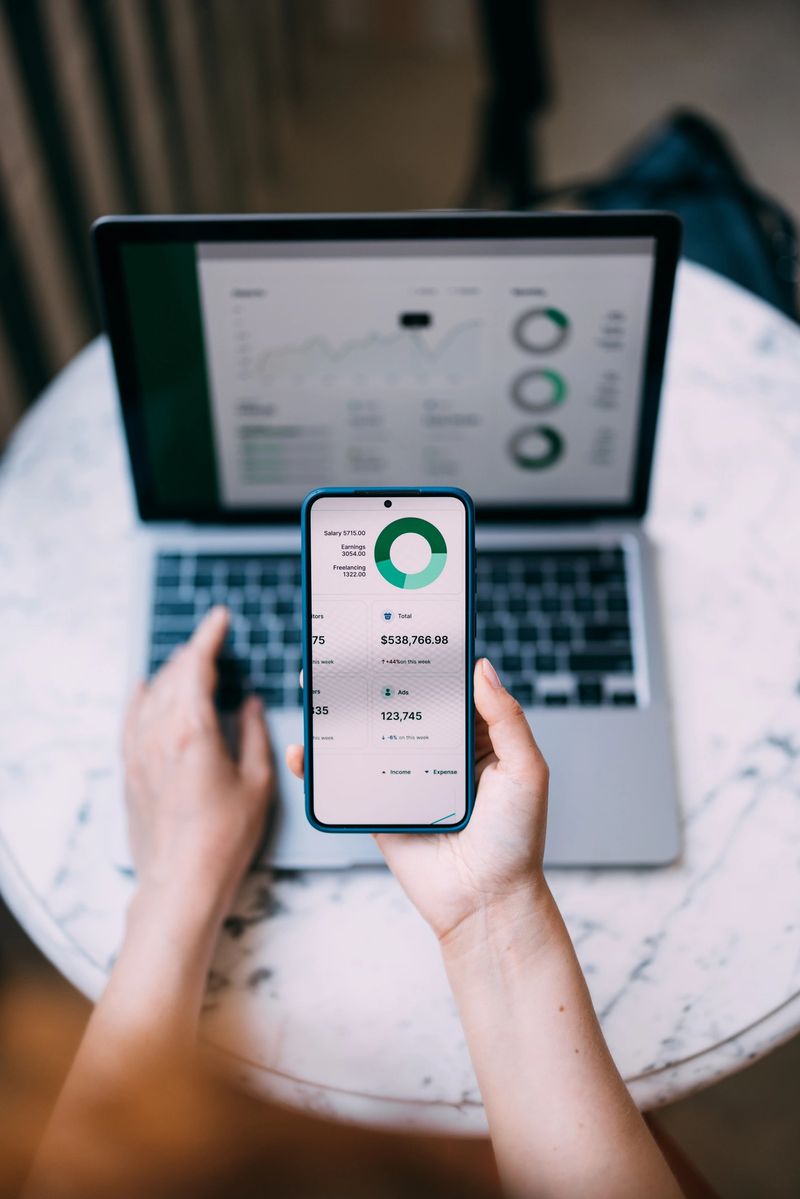 A person using a smartphone and laptop to review and analyze financial data with charts and graphs, illustrating modern technology, productivity, and financial management.