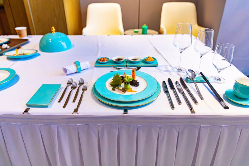 A meticulously plated dish showcases a rich combination of ingredients and five distinct colors: vibrant green leafy vegetables, crimson shrimp, golden sauce, white fish, and jet-black sea cucumber, accented with decorative garnishes. The vivid turquoise plate accentuates the colorful visual impact, highlighting the creative allure of refined dining.