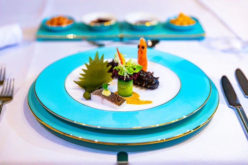A meticulously plated dish showcases a rich combination of ingredients and five distinct colors: vibrant green leafy vegetables, crimson shrimp, golden sauce, white fish, and jet-black sea cucumber, accented with decorative garnishes. The vivid turquoise plate accentuates the colorful visual impact, highlighting the creative allure of refined dining.