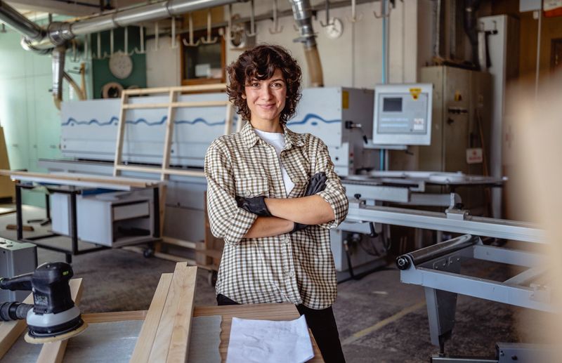 Une charpentière souriante dans un atelier avec des outils et des machines, portant une chemise à carreaux et des gants.