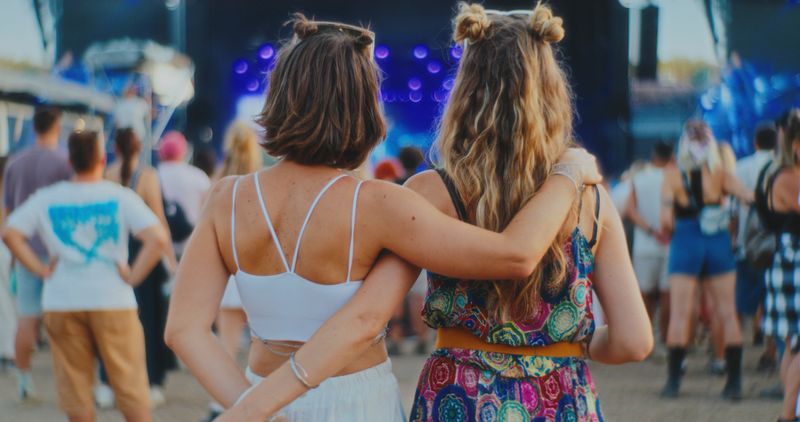 Two young Caucasian women are standing arm in arm at a lively music festival, featuring a colorful stage in the background. They are wearing casual summer outfits and are appearing happy and relaxed amidst the crowd.
