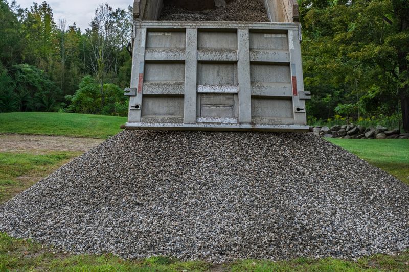 A dump truck unloads a large pile of crushed stone onto a grassy yard. The gravel will be used for home landscaping, driveways, or outdoor improvement projects. This image highlights bulk material delivery for residential or small-scale construction use.