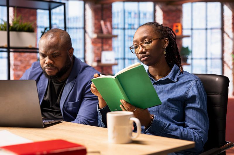 Black couple sitting together at home office and managing their affairs in their cozy loft. Young man solving work tasks on his laptop and his partner is reading a book for personal research.