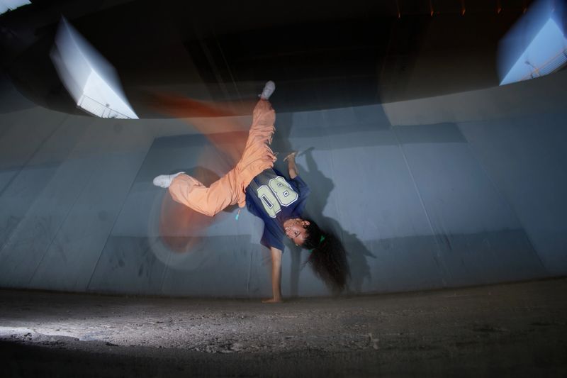 Girl performing dynamic one handed handstand breakdance move under urban overpass, long curly hair swinging, energetic movement captured mid air, Gen Z youth culture